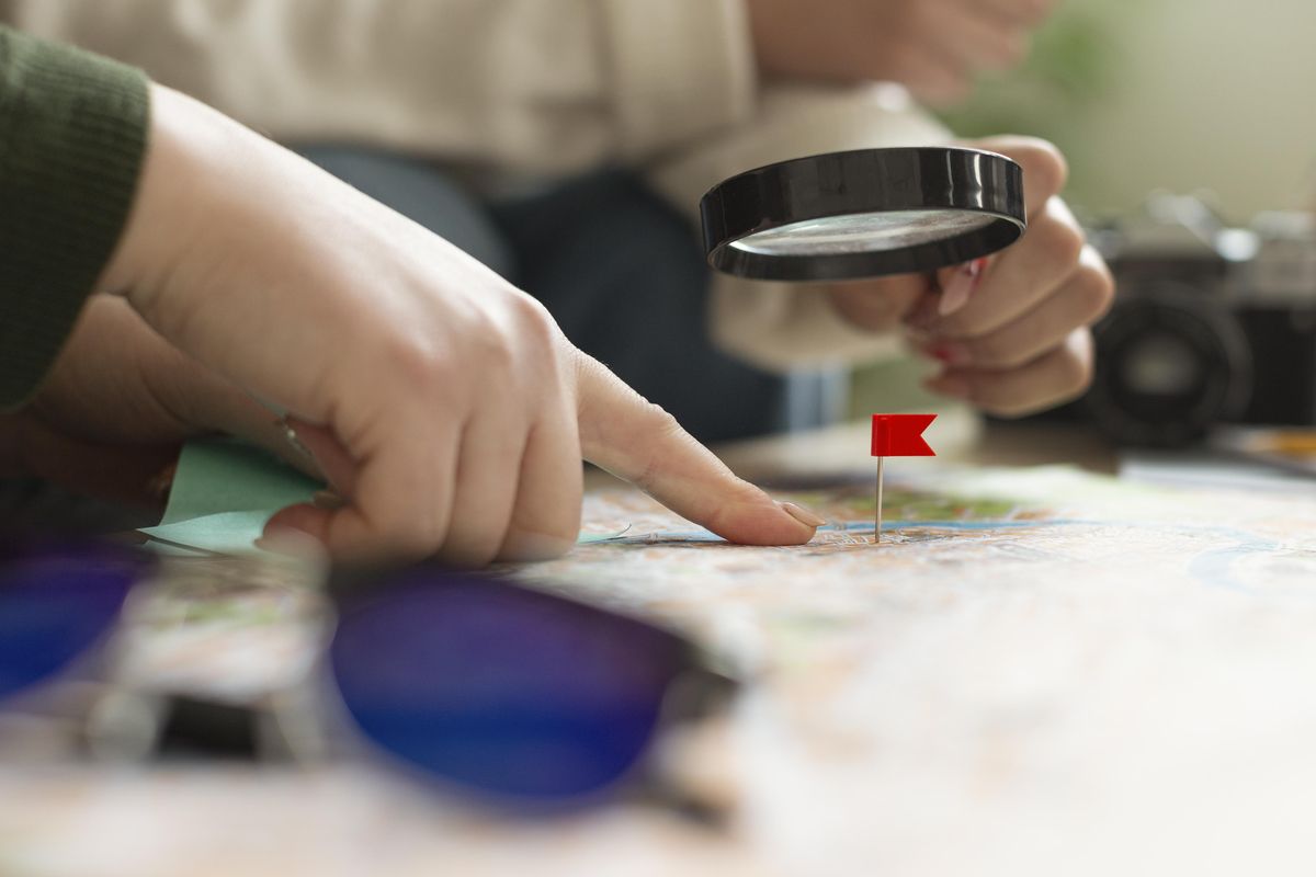 A finger and a magnifying glass over a map, with a red flag pinned on the target